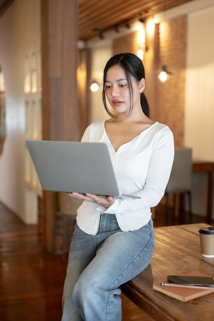 Une femme asiatique concentrée s'appuie sur une table et travaille sur son ordinateur portable dans un café