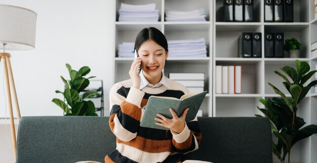 Femme asiatique confiante avec un sourire debout tenant un bloc-notes et une tablette dans le salon sur le canapé à la maison