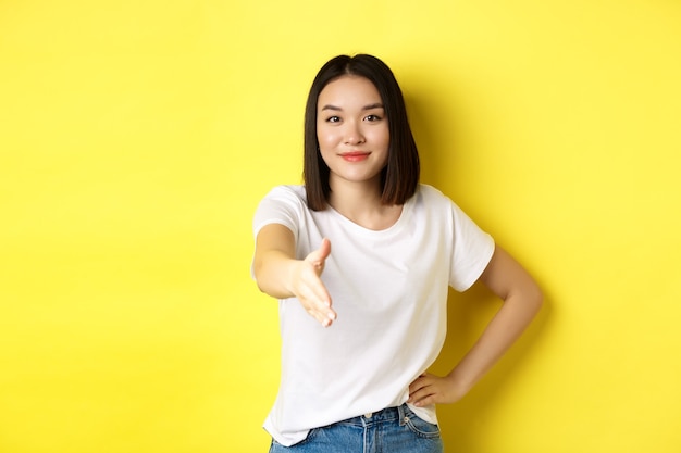 Femme asiatique confiante en t-shirt blanc, tendre la main pour la poignée de main et le geste de salutation, dire bonjour, se présenter, debout sur le jaune.