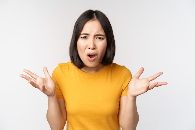 Une femme asiatique confuse et frustrée ne comprend pas qch qui a l'air désemparée en haussant les épaules debout en t-shirt jaune sur fond blanc