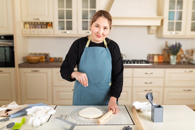 Femme asiatique cuisiner en tablier regarder la caméra saupoudrer de farine sur la pâte à pâtisserie de blé cuite sur un tapis de cuisson en silicone dans la cuisine moderne