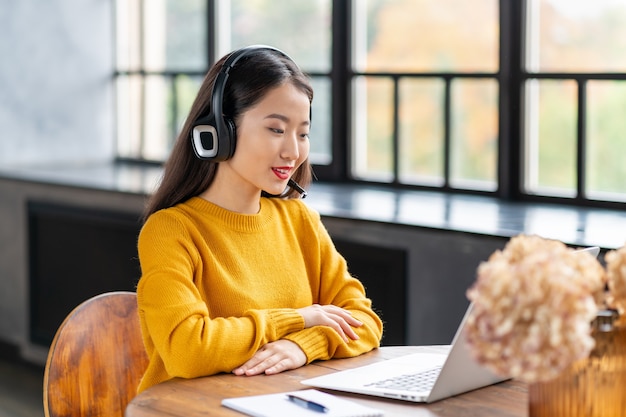 Femme asiatique dans le casque parlant par conférence téléphonique et chat vidéo sur ordinateur portable au bureau