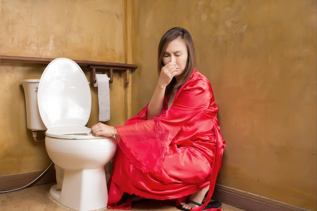 Femme asiatique dans une chemise de nuit en satin rouge nettoyant les toilettes à chasse d'eau la nuit parce que ça sent mauvais