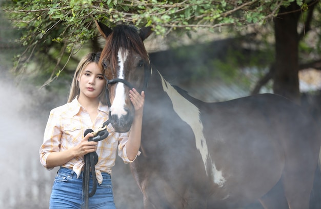 Femme asiatique dans un costume de cow-girl se tient avec un cheval dans une ferme d'élevage.