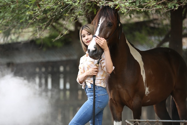 Femme asiatique dans un costume de cow-girl se tient avec un cheval dans une ferme d'élevage.