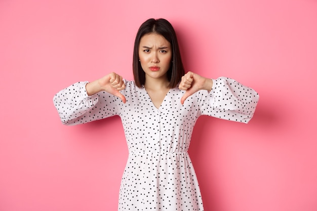 Femme asiatique déçue montrant le pouce vers le bas, désapprouvant et n'aimant pas quelque chose, montrant un jugement négatif, debout sur fond rose