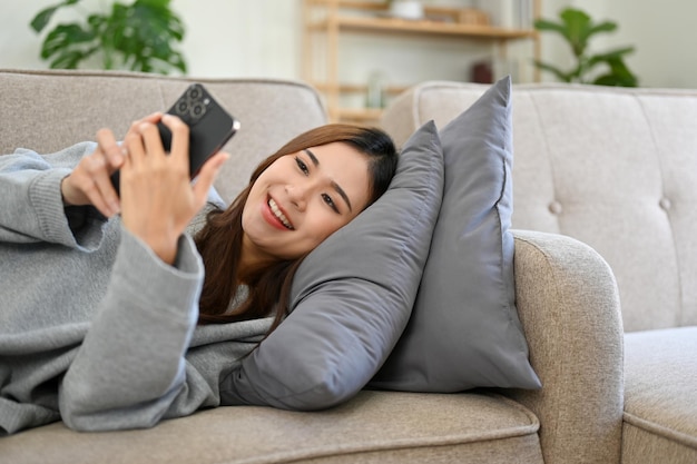 Femme Asiatique Détendue Et Heureuse Utilisant Un Smartphone Allongée Sur Son Canapé Confortable