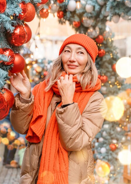 Femme asiatique en écharpe orange et chapeau marchant sur le marché de Noël décoré de lumières de Noël