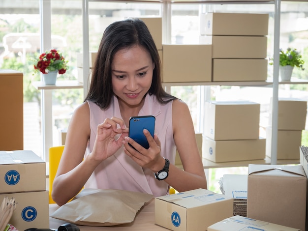 Femme asiatique écrit l'adresse sur la boîte de colis et vérifie la commande de produits sur smartphone