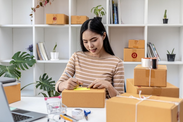 Femme asiatique écrivant les informations d'expédition du client sur la boîte à colis, elle possède une boutique en ligne, elle expédie des produits aux clients par l'intermédiaire d'une entreprise de messagerie privée. Concept de vente en ligne.