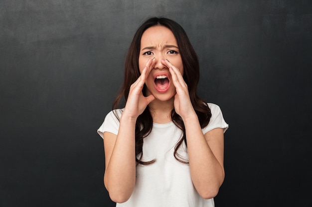 Femme asiatique émotionnelle en t-shirt décontracté hurlant ou appelant avec anxiété mettant les mains à la bouche, isolé sur un mur gris foncé