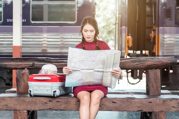 Femme asiatique enceinte en robe rouge portant des bagages rouges et regarde la carte à la gare traveltraveler avec sac à dos en concept de vacances d'été Thaïlande