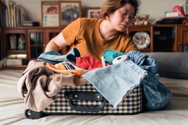 Une femme asiatique essaie de tout ranger dans les bagages avec des effets personnels en difficulté tels que des lunettes de soleil, un sac cosmétique pour appareil photo polaroid et un petit sac Emballage pour partir en vacances