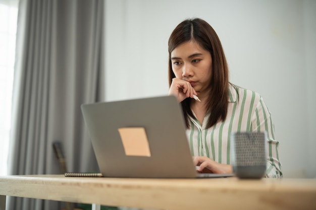 Une femme asiatique est assise à un bureau avec un ordinateur portable ouvert devant elle elle regarde l'écran et elle est profondément en pensée