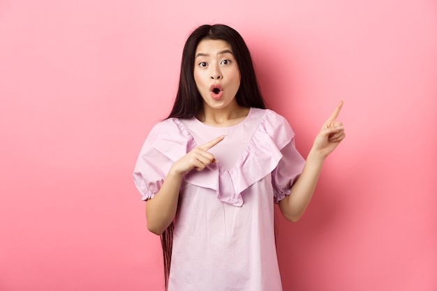 Une femme asiatique excitée a l'air étonnée, pointant vers la droite et haletante fascinée, vérifiant la publicité, debout sur fond rose.