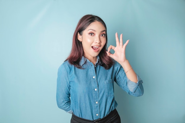 Femme asiatique excitée portant une chemise bleue donnant un geste de la main OK isolé par un fond bleu
