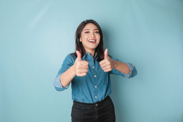 Une femme asiatique excitée portant une chemise bleue donne le pouce en l'air geste d'approbation isolé par fond bleu