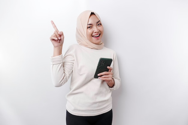 Femme asiatique excitée portant le hijab pointant vers l'espace de copie au-dessus d'elle tout en tenant son téléphone isolé sur fond blanc