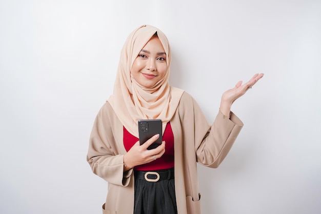 Femme asiatique excitée portant le hijab pointant vers l'espace de copie à côté d'elle tout en tenant son téléphone isolé sur fond blanc