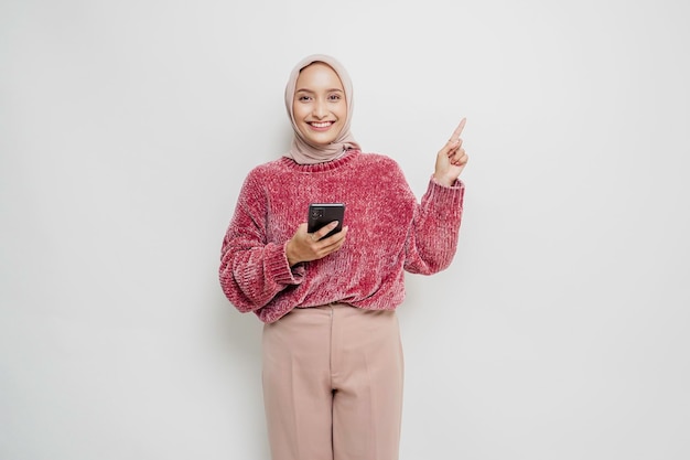 Femme asiatique excitée portant un pull rose et un hijab pointant vers l'espace de copie au-dessus d'elle tout en tenant son téléphone isolé sur fond blanc