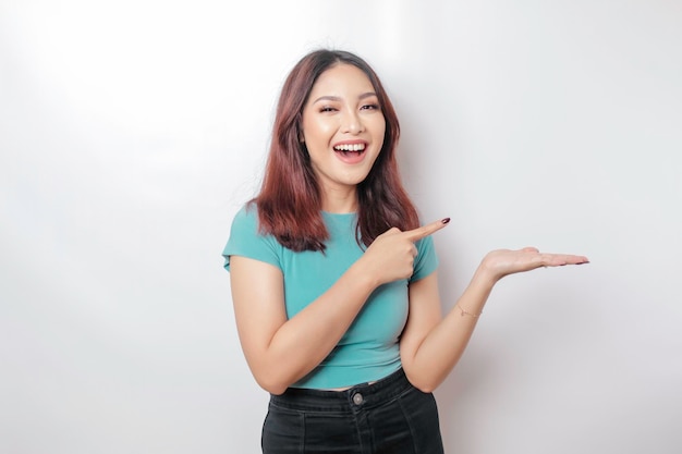 Femme asiatique excitée portant un t-shirt bleu pointant vers l'espace de copie à côté d'elle isolée par fond blanc