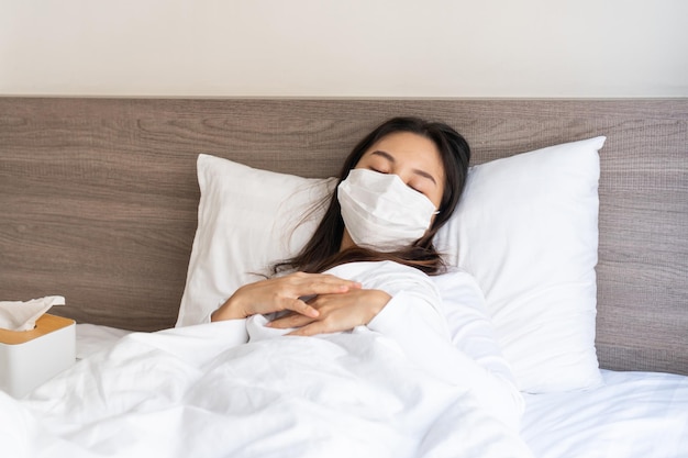 Une femme asiatique a une forte fièvre ou est infectée par le COVID19, reste à la maison et s'auto-met en quarantaine pendant la pandémie de coronavirus Une fille malade xAportant un masque facial a mal à la tête et tousse allongée sur le lit