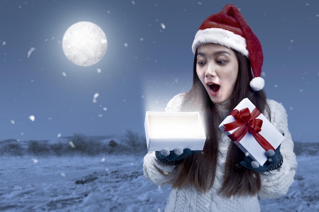 Femme asiatique avec des gants d'hiver et un bonnet de Noel ouvrant une boîte-cadeau blanche sur le champ de neige