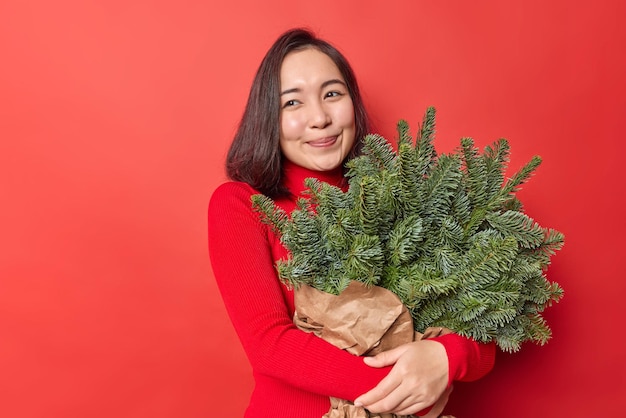 Une femme asiatique heureuse embrasse un gros bouquet de branches d'épinette verte enveloppées dans du papier rend le bouquet créatif de sapin semble rêveur pose sur un fond rouge vif rappelle les vacances passées