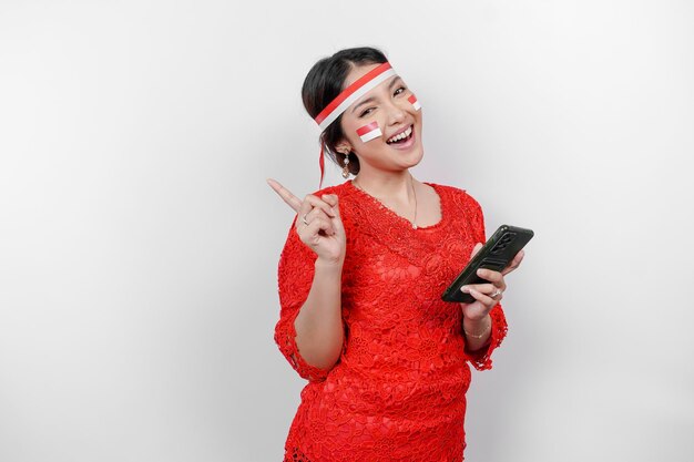 Photo une femme asiatique heureuse portant du kebaya rouge et un bandeau tenant son téléphone et pointant l'espace de copie au-dessus d'elle isolée par fond blanc le jour de l'indépendance de l'indonésie