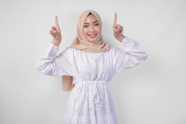 Femme asiatique heureuse portant un hijab et une robe blanche pointant vers l'espace de copie au-dessus debout sur un fond blanc isolé concept publicitaire Ramadan et Eid Mubarak