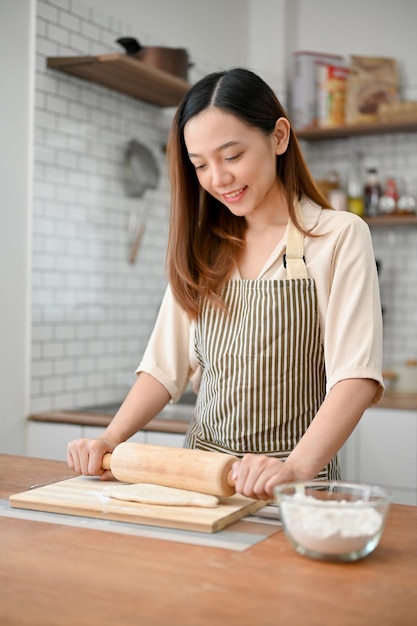 Une femme asiatique heureuse en tablier pétrit de la pâte crue avec un rouleau à pâtisserie faisant de la pâtisserie dans la cuisine