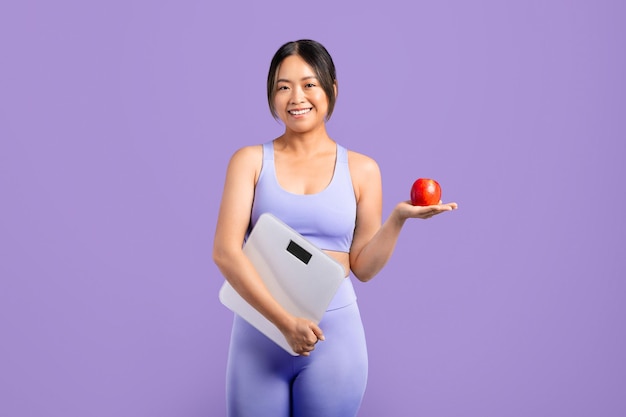 Une femme asiatique heureuse en vêtements de sport tenant une balance et une pomme rouge.