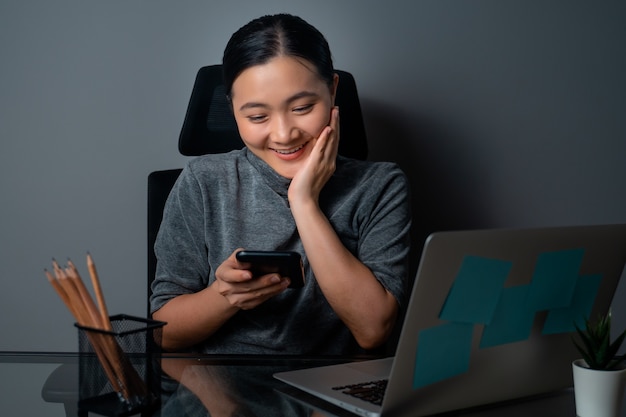 Femme asiatique heureux souriant à l'aide de téléphone intelligent et travaillant sur un ordinateur portable au bureau