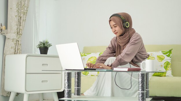 Femme asiatique en hijab travaillant à domicile en tapant un ordinateur portable tout en écoutant