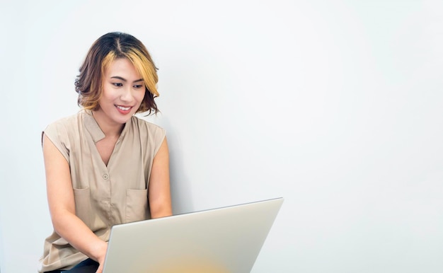 Femme asiatique indépendante avec des cheveux courts à la mode en regardant l'écran tout en travaillant avec un ordinateur portable sur fond de mur blanc avec espace de copie