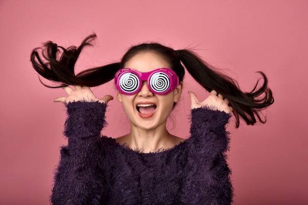 Femme asiatique joyeuse, fille excitée portant des lunettes drôles sur fond rose isolé, fille joyeuse sautant avec bonheur.