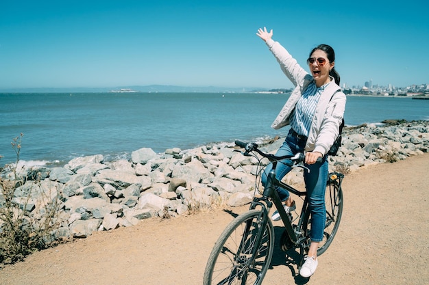 une femme asiatique joyeuse lève le bras et crie de joie à distance à vélo tout en visitant la station balnéaire de la marina lors de vacances d'été ensoleillées en Californie.