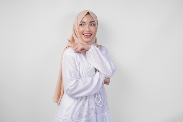Une femme asiatique joyeuse portant un hijab et une robe blanche pointant vers l'espace de copie derrière debout sur un fond blanc isolé concept publicitaire Ramadan et Eid Mubarak