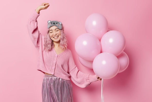 Une femme asiatique joyeuse et positive danse sans soucis garde le bras levé tient un tas de ballons gonflés porte un pull décontracté et une jupe plissée un masque de sommeil sur le front profite de la fête Événement Fesitve