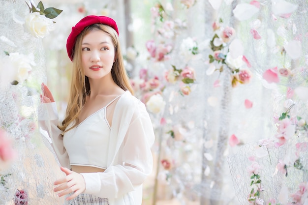 Une femme asiatique a de longs cheveux bronze et porte une robe blanche à bonnet rouge alors qu'elle se tient au milieu d'un jardin fleuri