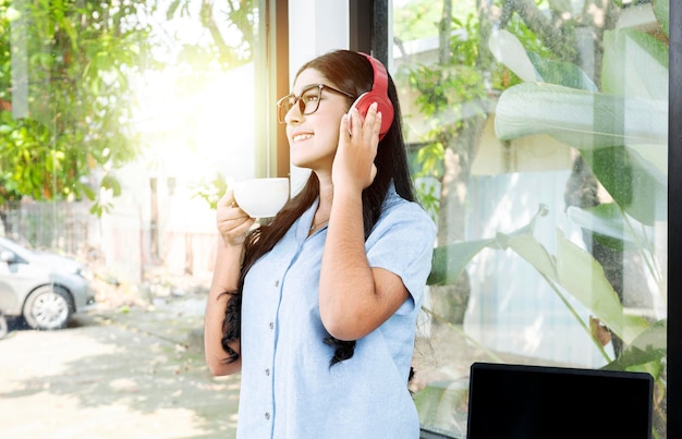 Femme asiatique avec des lunettes écoutant de la musique avec des écouteurs et tenant une tasse de café avec un ordinateur portable sur la table