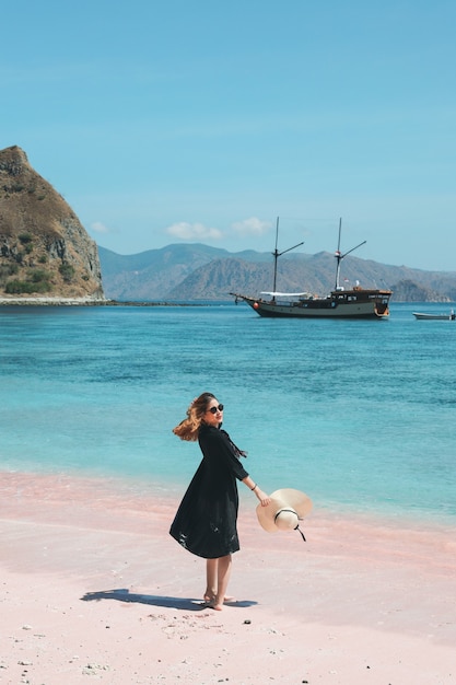 Femme asiatique à lunettes de soleil tenant un chapeau d'été profitant de vacances sur une plage de sable rose avec un bateau