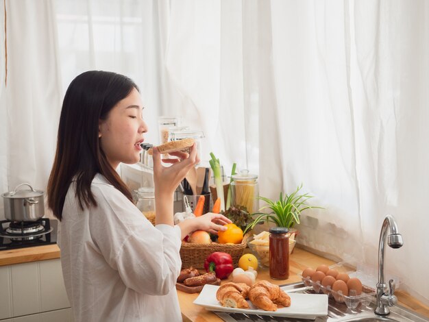 Femme asiatique mangeant du pain tout en préparant un repas dans la cuisine