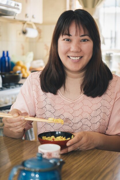 Une femme asiatique mangeant du riz avec des baguettes regarde et sourit à la caméra.