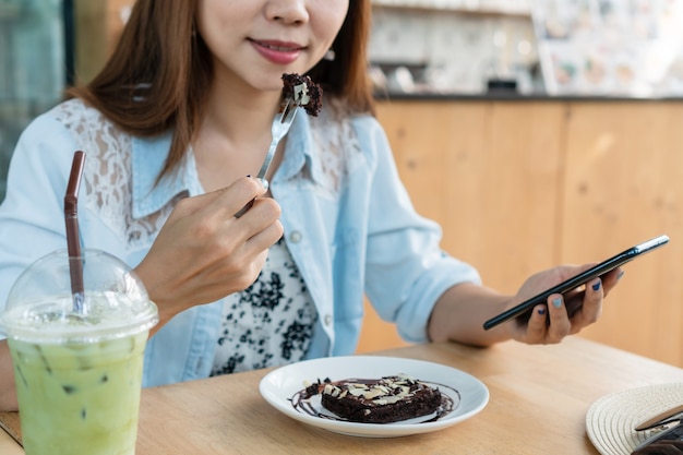 Femme asiatique, manger un gâteau brownie tout en utilisant un smartphone.