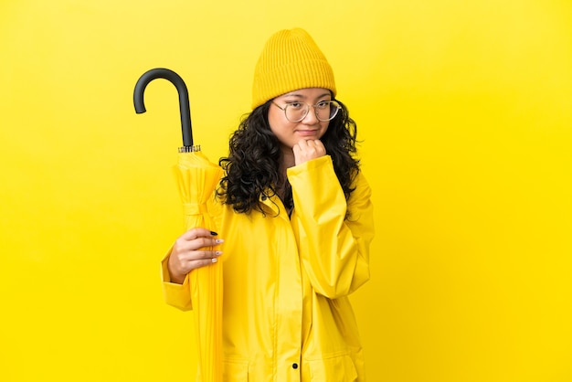 Femme asiatique avec manteau imperméable et parapluie isolé sur fond jaune ayant des doutes