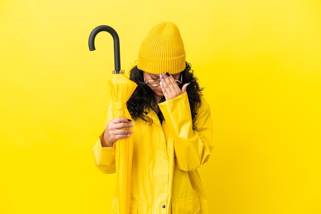 Femme asiatique avec manteau imperméable et parapluie isolé sur fond jaune avec une expression fatiguée et malade