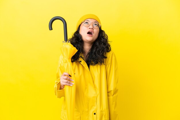 Femme asiatique avec manteau imperméable et parapluie isolé sur fond jaune en levant et avec une expression surprise