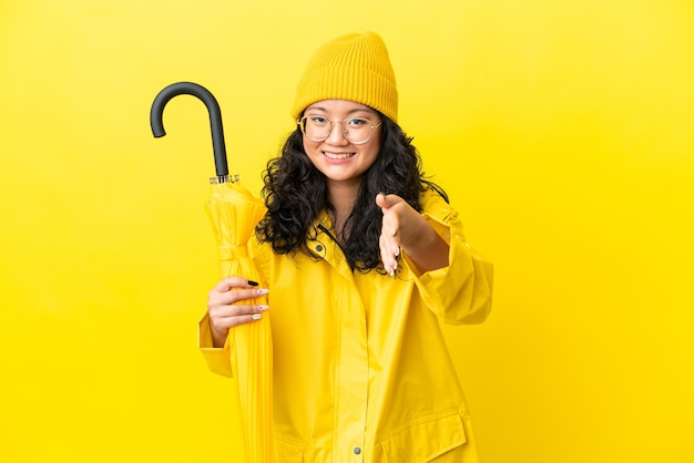 Femme asiatique avec manteau imperméable et parapluie isolé sur fond jaune se serrant la main pour conclure une bonne affaire