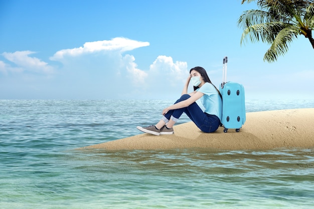 Femme asiatique avec un masque facial assis avec une valise sur la plage